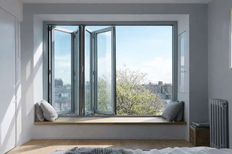Bright modern bedroom with black-framed folding windows, a built-in window seat, soft pillows, and a city view through the open glass panels.