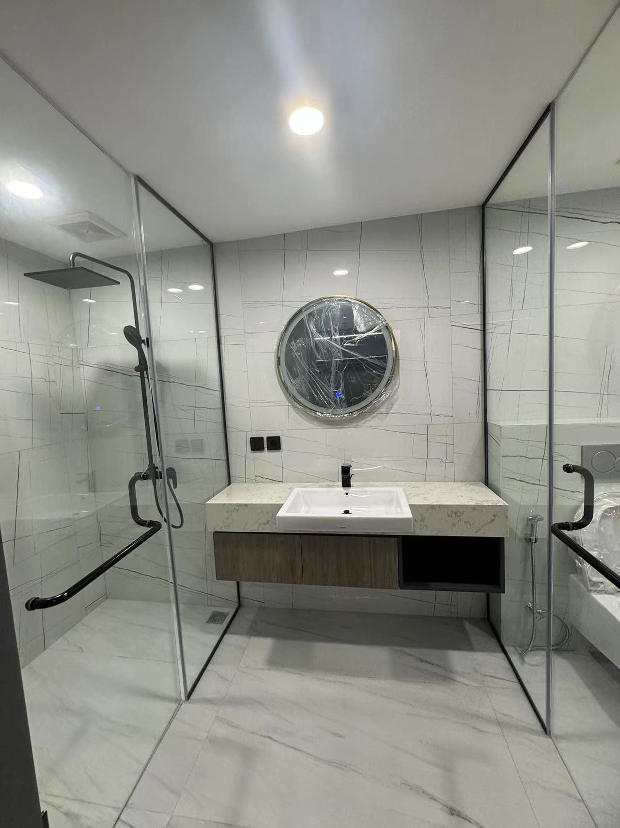 Medan Manhattan Apartment Renovation in Indonesia 13 Modern bathroom with floating vanity, glass shower partition, marble-look wall tiles, and black fixtures.