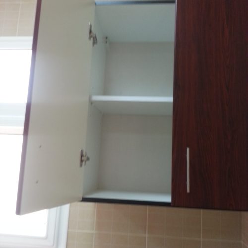 In this Abu Dhabi apartment whole house custom project, A cabinet with a cherry wood veneer and silver metal handles, featuring a white interior, installed next to a window, offering a stylish and functional storage solution.